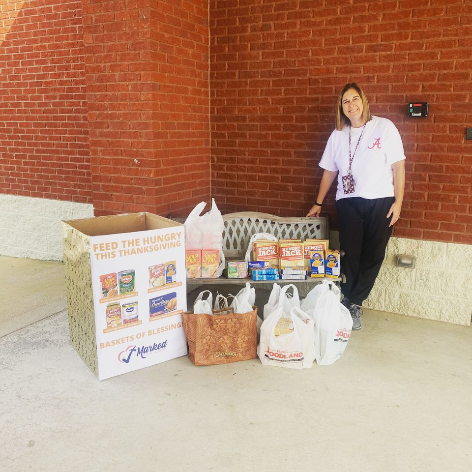 A smiling woman stands beside a Baskets of Blessings collection box and a bench full of donated stuffing, mashed potatoes, and macaroni and cheese.