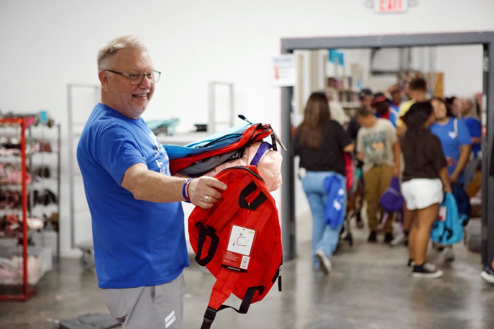A smiling volunteer carries an armful of new red, blue, and pink backpacks while families line up behind him.