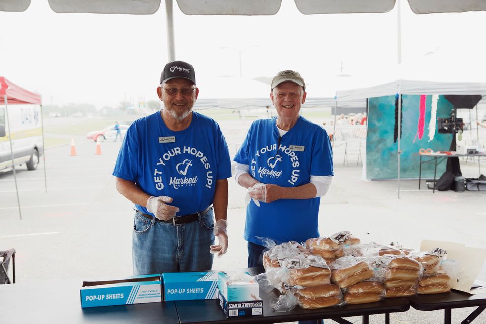 Lawrence and Ronnie, volunteers in blue Get Your Hopes Up shirts and food-service gloves, stand behind a table of hamburger buns under a tent.