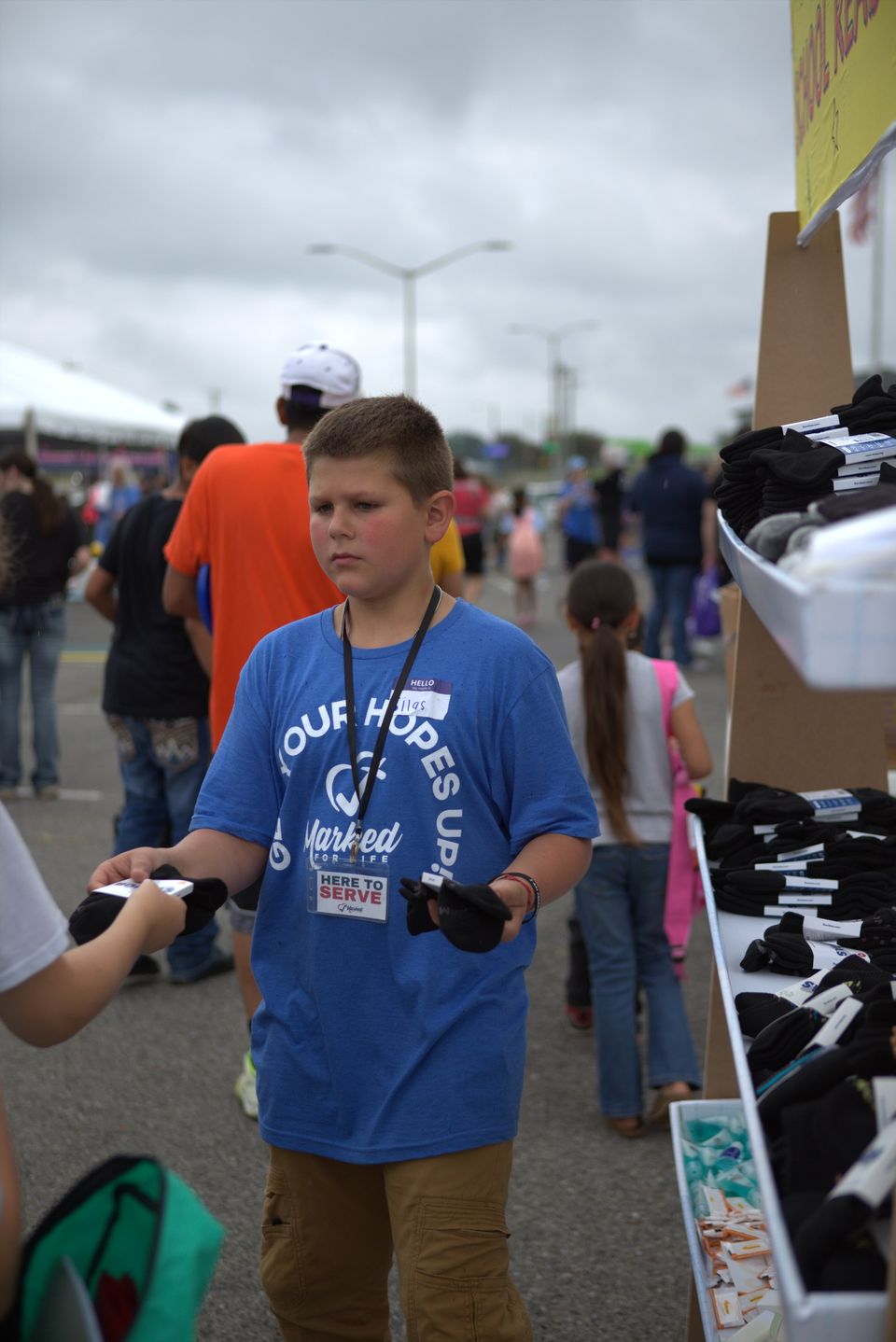 A boy in a Marked For Life shirt carries supplies past a giveaway table under an overcast sky.