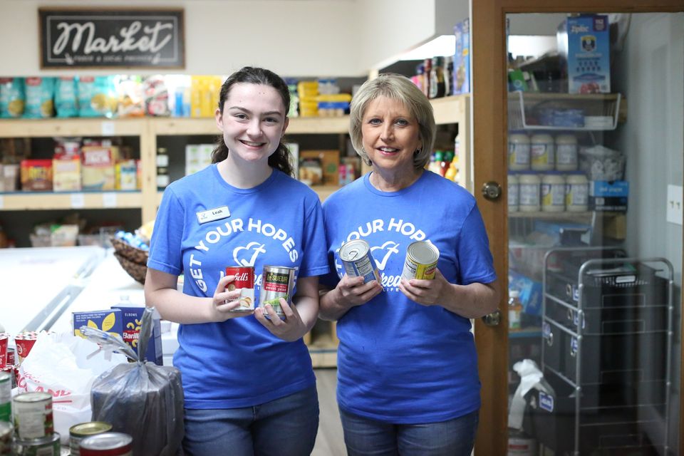 Leah and a fellow volunteer hold up canned goods in the Hope Center Market under the chalkboard Market sign.