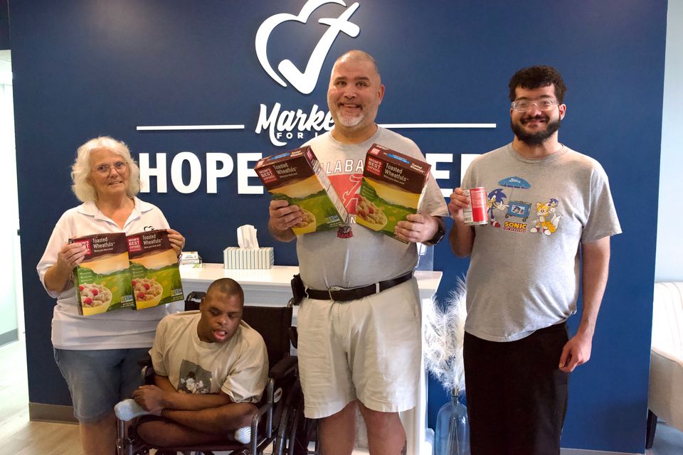 Four neighbors, one in a wheelchair, hold up boxes of cereal in front of the Hope Center wall sign.