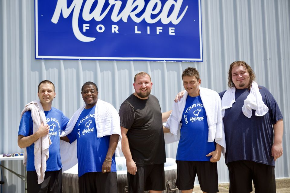 Five men with towels around their necks smile under the Marked For Life sign after being baptized.