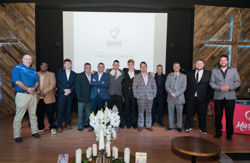 Hope House students and staff in suits line up on stage in front of a lit wooden cross at a Hope House graduation.
