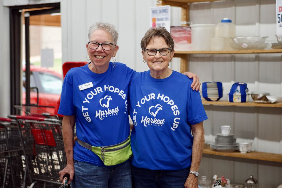 Diane and a fellow volunteer in blue Get Your Hopes Up shirts smile beside shelves of housewares at the Thrift Store.