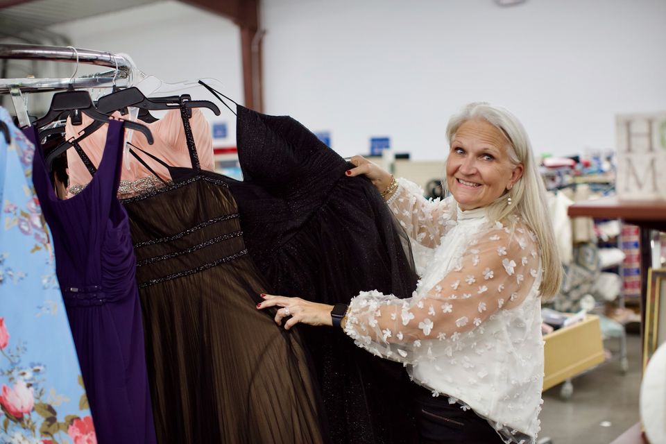 A volunteer with silver hair holds up a black gown from a rack of formal dresses at the Marked For Life Thrift Store.