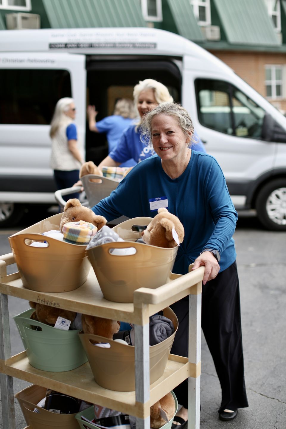 Julie pushes a cart of baskets filled with teddy bears and blankets while volunteers unload a church van behind her.