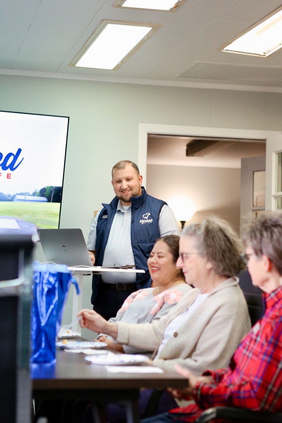 Logan LeGrande stands at a laptop in a Marked For Life vest while three new volunteers laugh at the table during orientation.