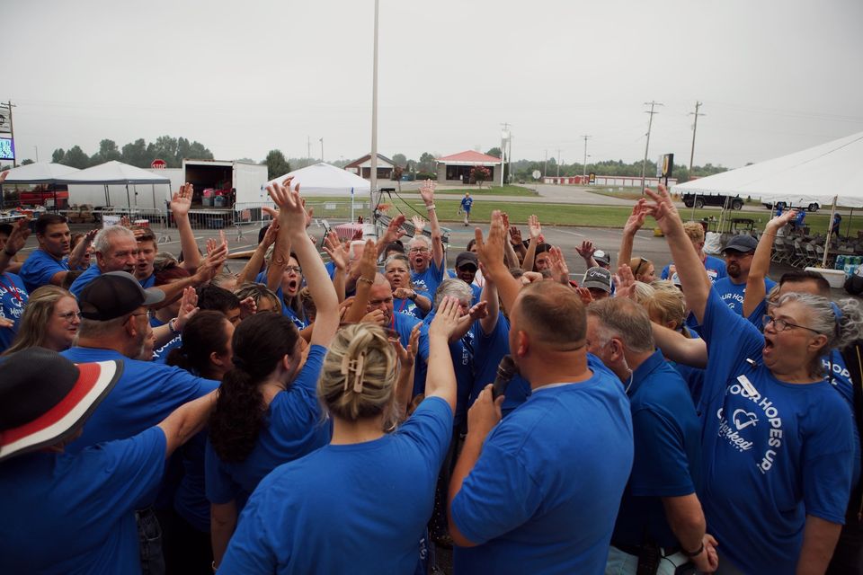 Dozens of volunteers in blue Get Your Hopes Up shirts raise their hands in a huddle while Logan speaks into a microphone.