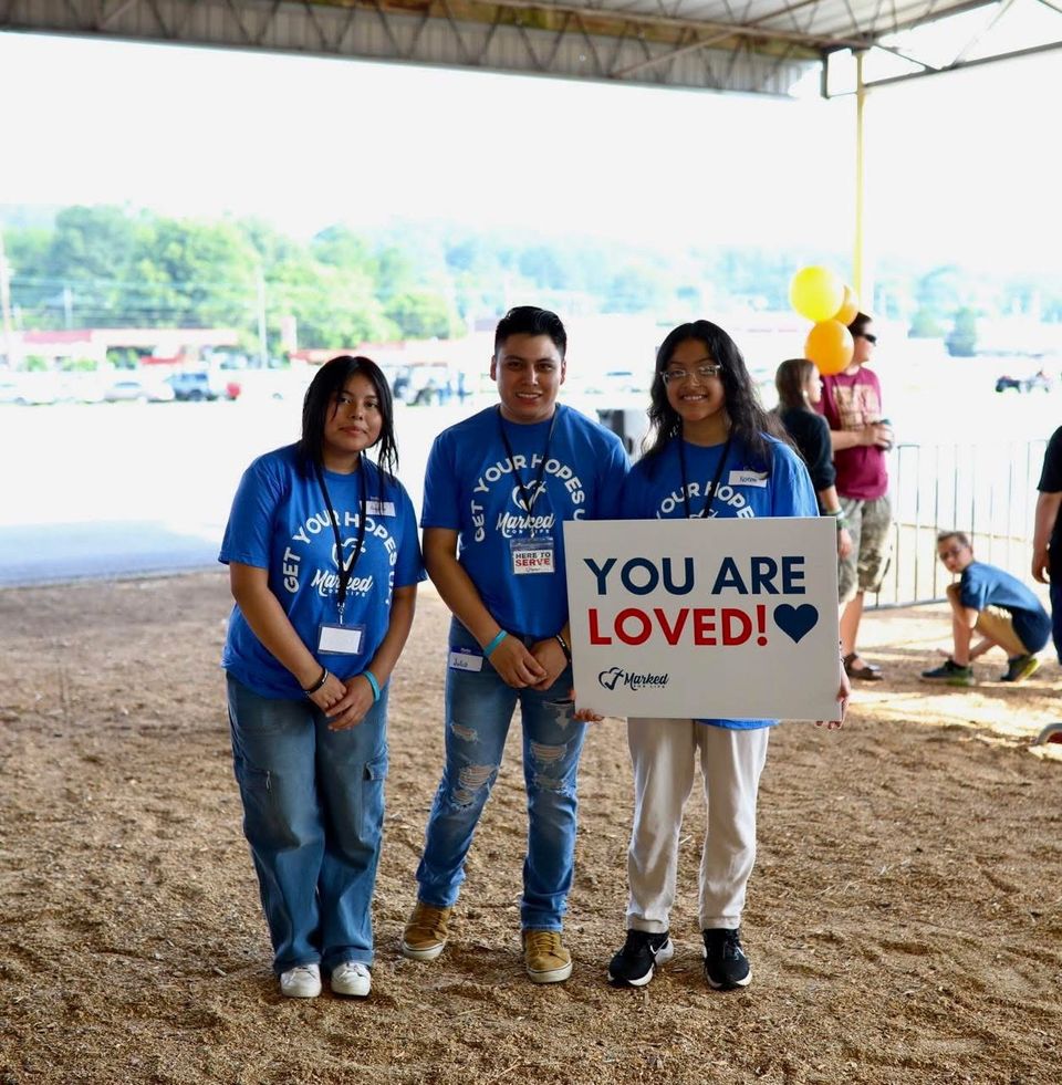 Three teen volunteers in blue shirts hold a sign reading You Are Loved under a pavilion.